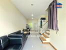 Living room with black sofa, dining area, staircase, and a website URL on the wall.