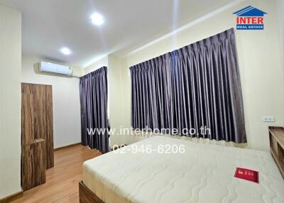 Spacious, well-lit bedroom with wooden flooring, large windows with grey curtains, air conditioning, and a comfortable bed