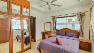 Well-lit bedroom with purple bedding, wooden furniture, and ceiling fan