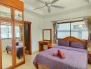 Well-lit bedroom with purple bedding, wooden furniture, and ceiling fan