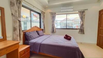 A bright bedroom with purple bedding, wooden furniture, and two large windows.