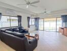 Spacious living room with black leather sofa, glass coffee table, wooden cabinets, ceiling fans, air conditioning units, and large windows with blue curtains