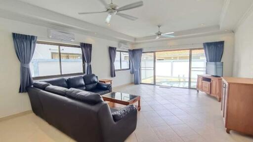 Spacious living room with black leather sofa, glass coffee table, wooden cabinets, ceiling fans, air conditioning units, and large windows with blue curtains