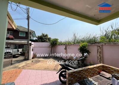 Outdoor space with tiled flooring and a cover, featuring a motorcycle, nearby buildings, and greenery