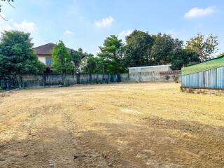 Spacious outdoor lot with bare ground, trees, and buildings in the background