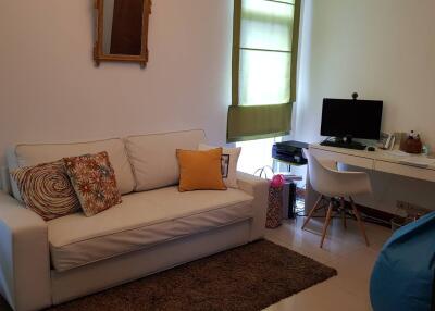 A cozy living room with a white sofa, decorative pillows, a desk with a computer, and a window with green blinds