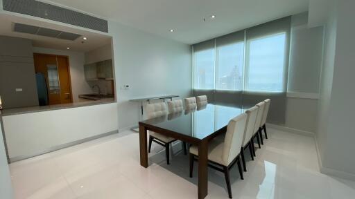 Modern dining room with a glass-top table and eight white upholstered chairs, adjacent to an open kitchen