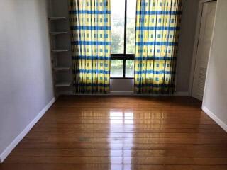 Empty room with hardwood flooring, window with patterned curtains, and built-in shelving