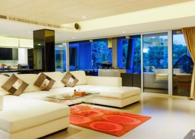 Modern living room with large white sectional sofa, glass coffee table, red rug, and sliding glass doors leading to a balcony with a view of the forest