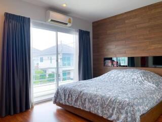 A cozy bedroom with wooden flooring, a large bed with grey floral bedding, a wooden accent wall, and sliding glass doors opening to a balcony with a view.