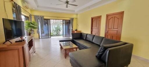 Bright and spacious living room with black sectional sofa, glass coffee table, large TV stand, ceiling fan, and large sliding glass doors leading to the garden.