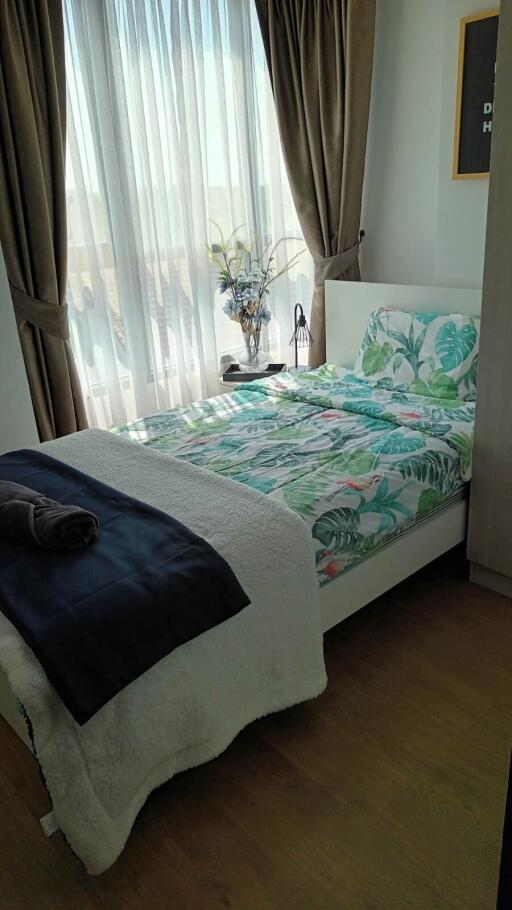 A cozy bedroom with a large window, floral bedding, and a side table with a decorative plant.