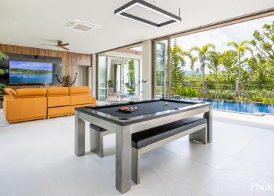 Modern living area with pool table and large windows overlooking a pool and tropical scenery