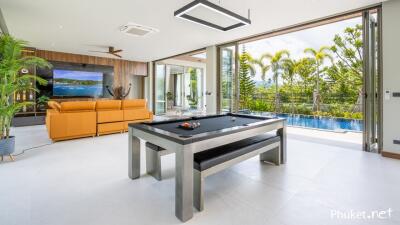 Modern living area with pool table and large windows overlooking a pool and tropical scenery