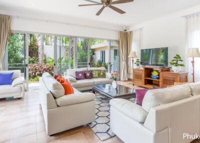 Spacious living room with large windows, white sofas, and a ceiling fan.