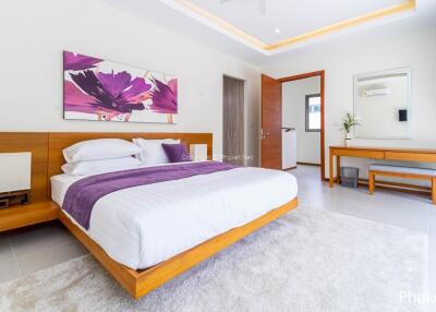 Modern bedroom with wooden furniture, white and purple bedding, decorative artwork, and a desk next to a large window with curtains