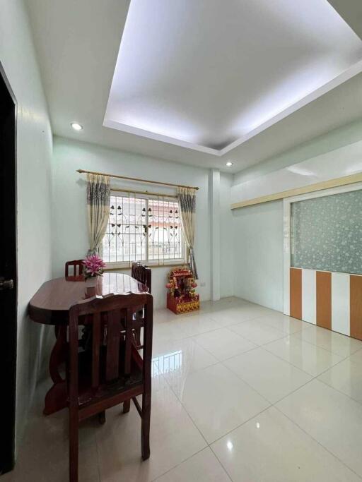 Bright dining area with wooden chairs and a table, decorative ceiling, tiled floor, window with curtains, and a partially partitioned wall.