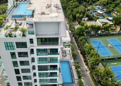 Aerial view of a modern multi-story building with multiple pools and tennis courts nearby.