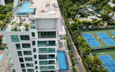 Aerial view of a modern multi-story building with multiple pools and tennis courts nearby.