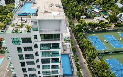 Aerial view of a modern multi-story building with multiple pools and tennis courts nearby.