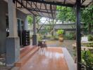 Covered outdoor patio area with a small table and chairs, providing a view of a lush garden.