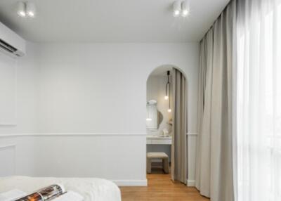 Modern bedroom with an arched doorway leading to a vanity area, hardwood flooring, large window with curtains, and a ceiling-mounted air conditioning unit.