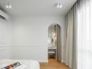 Modern bedroom with an arched doorway leading to a vanity area, hardwood flooring, large window with curtains, and a ceiling-mounted air conditioning unit.