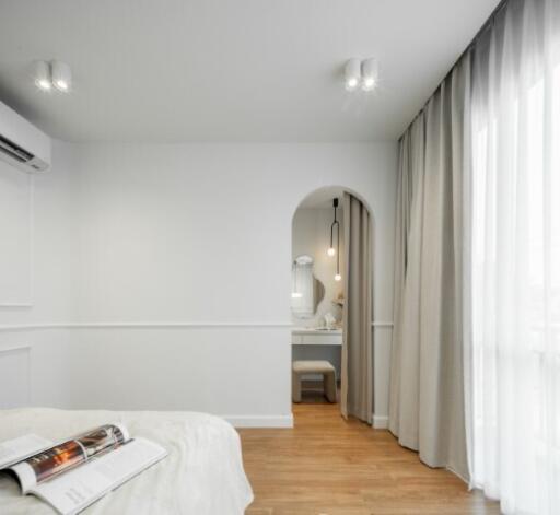 Modern bedroom with an arched doorway leading to a vanity area, hardwood flooring, large window with curtains, and a ceiling-mounted air conditioning unit.