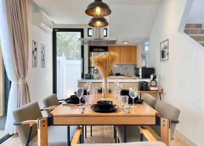 A well-lit dining room with a wooden dining table set for a meal, modern lighting fixtures, and an open kitchen in the background.