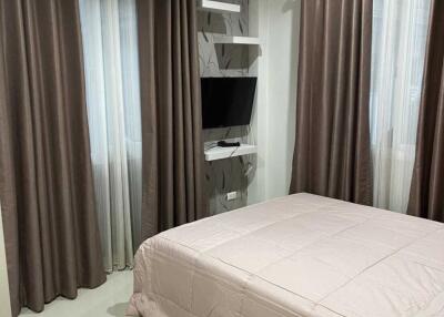 A modern bedroom with brown curtains, a wall-mounted TV, and a bed with a light-colored comforter.