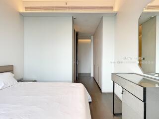 Modern bedroom with minimalistic decor, featuring a large bed with white linens, a sleek bedside table, a glass-topped desk, and an en-suite closet.