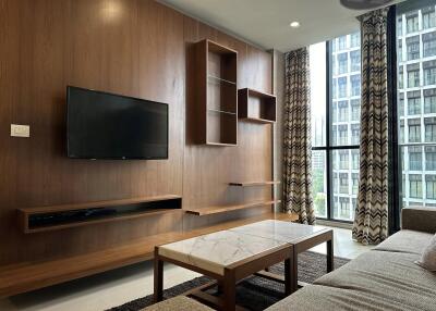 Modern living room with a wall-mounted TV and bookshelf, large windows with city view, and a coffee table