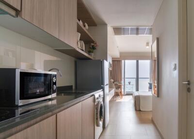 Modern kitchen area with wooden cabinets, microwave, washing machine, and view towards a living space with table and chair near large windows