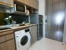 Compact kitchen with wooden cabinets, washing machine, microwave, fridge, and sink area
