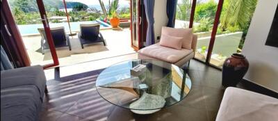 Modern living room with a glass coffee table, seating area, and large glass doors opening to a patio with a pool view.