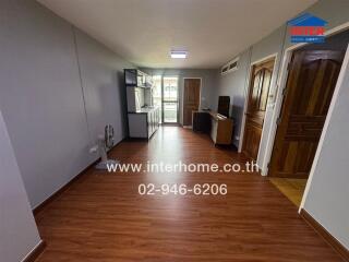 Spacious living area with wooden floor, connected to a kitchen, featuring light grey walls and wooden doors