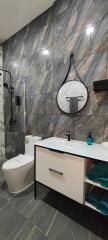 Modern bathroom with marble tile walls, a round mirror, a toilet, and a vanity with storage space