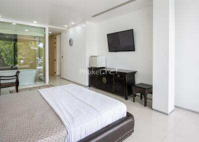 Spacious modern bedroom with large bed, wall-mounted TV, and office space with window view