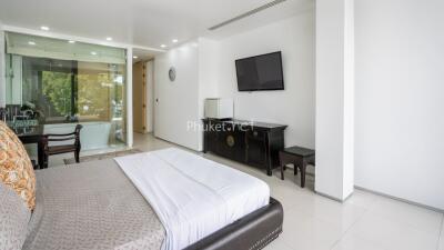 Spacious modern bedroom with large bed, wall-mounted TV, and office space with window view