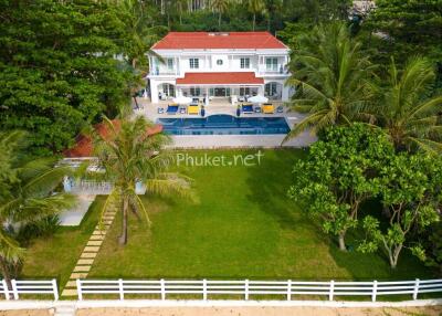 Exterior view of a large house with a red roof, balcony, and swimming pool surrounded by lush greenery