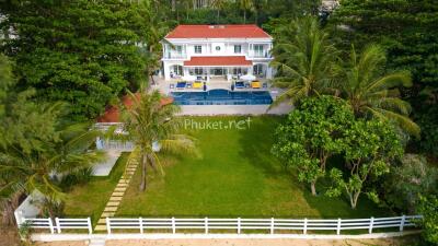 Exterior view of a large house with a red roof, balcony, and swimming pool surrounded by lush greenery