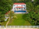 Exterior view of a large house with a red roof, balcony, and swimming pool surrounded by lush greenery