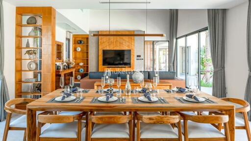 Modern dining room with wooden table and chairs, set up for a meal, connected to living area with TV and large windows.