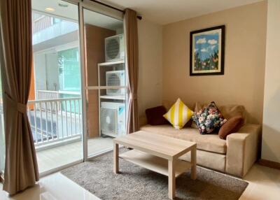 Cozy living room with brown sofa, decorative pillows, coffee table, and a framed artwork; adjacent to the balcony with large glass doors and air conditioning units outside.