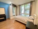 Bedroom with wooden flooring, blue accent wall, large window, bed, wardrobe, and dresser