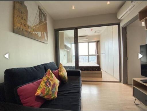 Modern living room with a view into the bedroom, featuring a black sofa with colorful pillows and a wall art