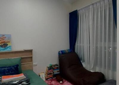 Bedroom with a green bedspread, brown lounge chair, books, and window with curtains