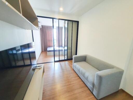 Modern living area with a grey couch and wooden flooring, separated by a glass partition from the bedroom