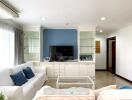 Well-lit living room with white sofas, blue accent wall, built-in storage, and flat-screen TV