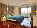 Bedroom with wooden floor, large bed, bedside tables, lamps, air conditioner, and balcony access.